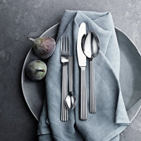 A plate with a folded gray cloth holds the Georg Jensen Bernadotte Flatware 5 Piece Setting—fork, knife, spoon, and teaspoon—with two figs beside the cutlery on a stone-like gray surface.