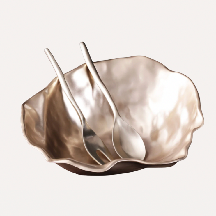 The Sierra Maia Salad Servers by Beatriz Ball feature two handmade, metallic utensils in silver or champagne tones, elegantly placed inside a shiny, irregularly shaped bowl against a light background.
