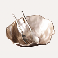 The Sierra Maia Salad Servers by Beatriz Ball feature two handmade, metallic utensils in silver or champagne tones, elegantly placed inside a shiny, irregularly shaped bowl against a light background.