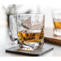 A Simon Pearce Woodbury Bourbon glass, textured and filled with amber whiskey, rests on a gray coaster. Another lead-free glass by Simon Pearce is blurred in the background on a wooden surface in a modern, well-lit setting.