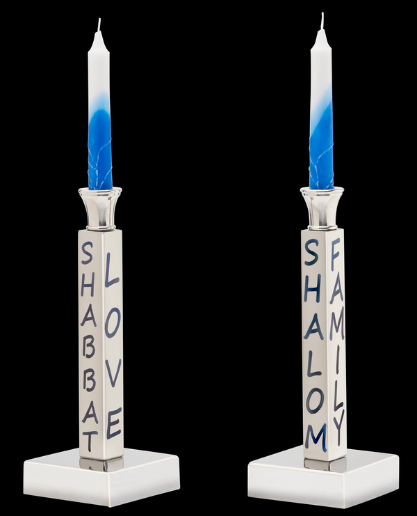 The Inspired Generations Shabbat Candlestick Set includes two white and blue candles in square silver holders, each engraved with "SHABBAT LOVE" or "SHALOM FAMILY," creating an elegant display against a black background.
