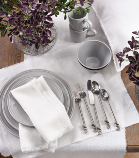 A table set with stacked gray plates, silverware, gray bowls and cups, vases with purple-green foliage, and Saro Trading’s Fringed Stone Washed Napkin Ivory Set of 4, all on a white tablecloth.