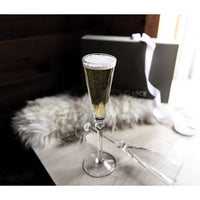 A Hartland Flute Set of 2 by Simon Pearce, filled with sparkling wine and placed beside an empty glass, a white throw, and a gray gift box with ribbon on a wooden table—an elegant choice for a chic wedding gift.