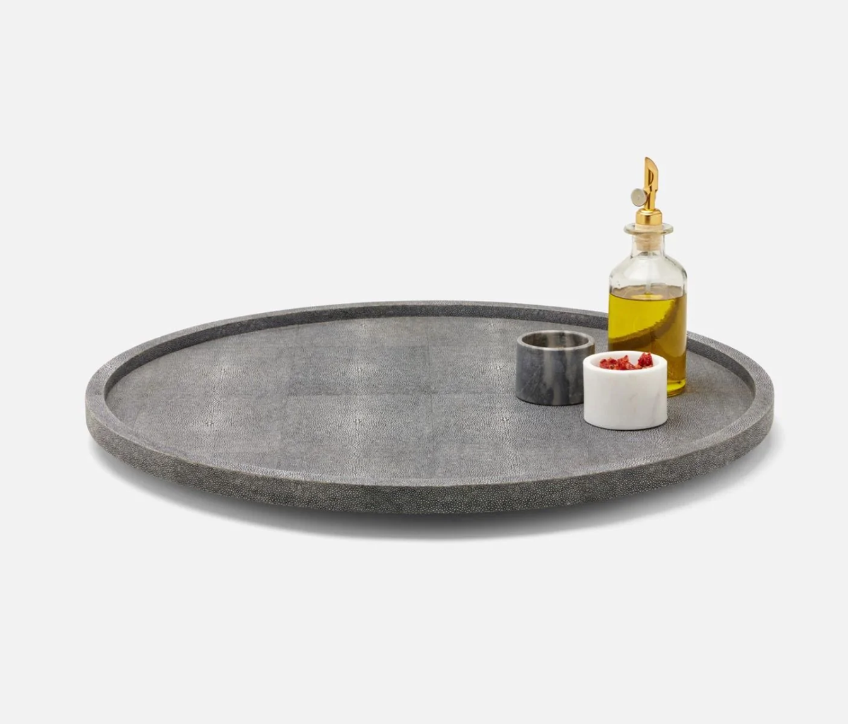 The Calvin Faux Shagreen Lazy Susan in Grey by Blue Pheasant displays a glass olive oil bottle with gold spout, a small gray marble cup, and a white marble bowl of red spices—creating an elegant centerpiece for entertaining.