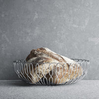 The Georg Jensen Alfredo Bread Basket Large offers a sleek stainless steel design, showcasing rustic bread beautifully on any gray stone surface—perfect for elevating your next dinner party.
