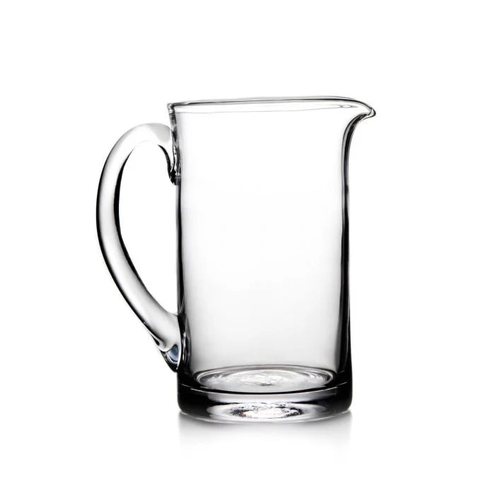 The Simon Pearce Ascutney Pitcher Medium is a handmade clear glass pitcher with a curved handle and spout, shown empty against a white background.