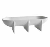 The FS Objects Standing Bowl Oval features a modern white oval design with three evenly spaced vertical support fins, serving as a sleek decorative centerpiece reminiscent of a contemporary fruit bowl.