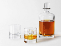The Fluted Decanter Large by Richard Brendon, filled with amber whiskey, and two mouth-blown crystal glasses—one with ice—are displayed against a white background.