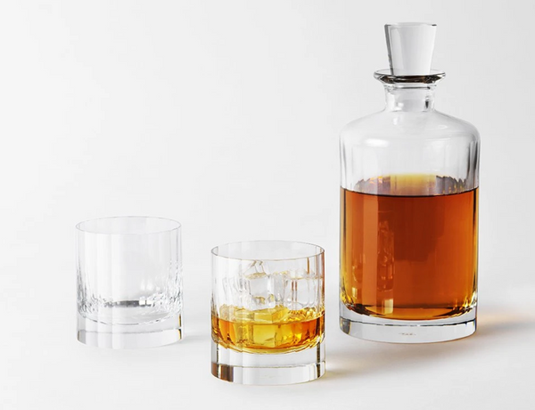 The Fluted Decanter Large by Richard Brendon, filled with amber whiskey, and two mouth-blown crystal glasses—one with ice—are displayed against a white background.