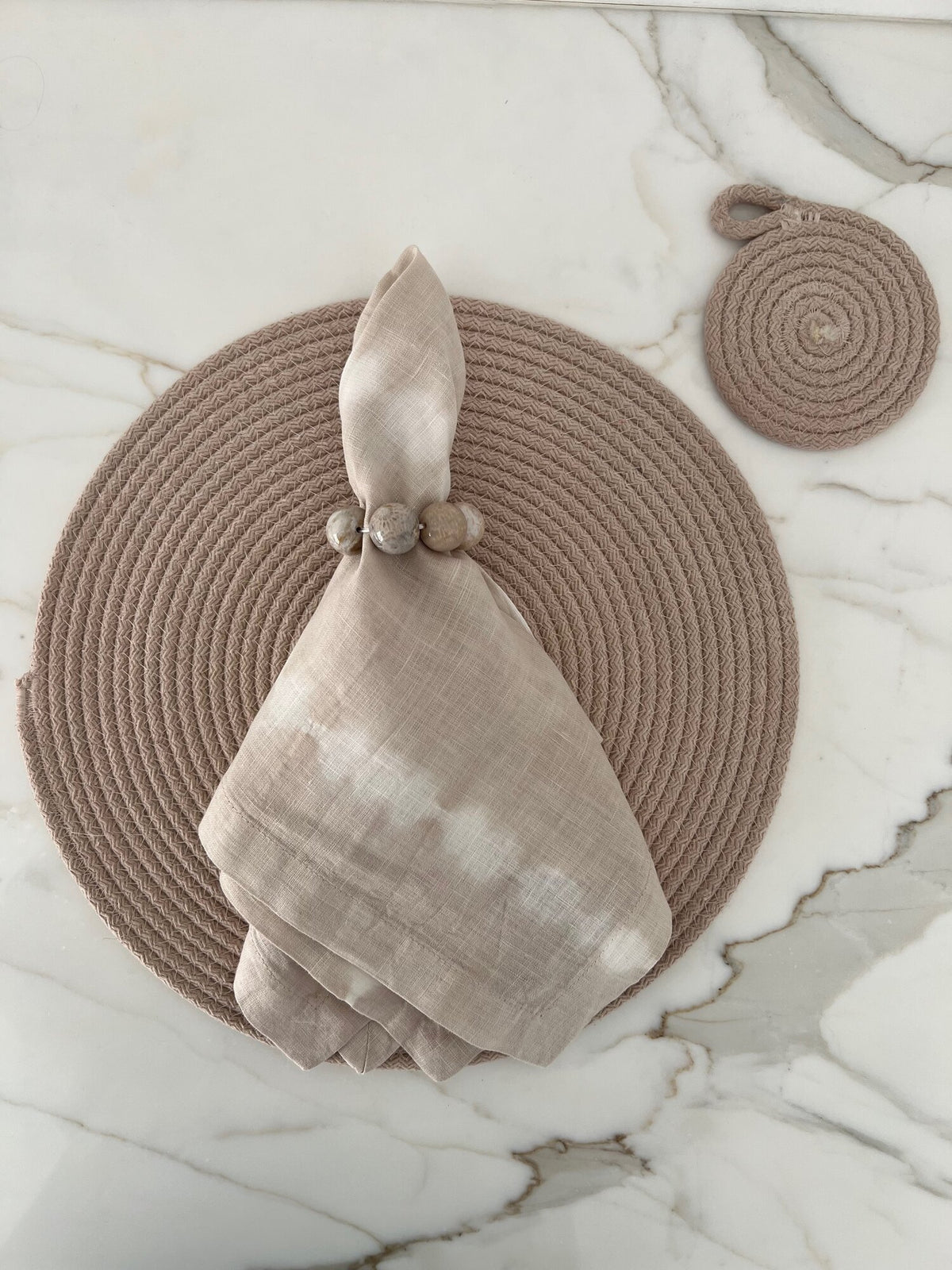 A Whigstock Studio Marble Bead Napkin Ring in cream secures a cloth napkin on a beige woven placemat atop marble, with a matching round coaster placed at the top right.