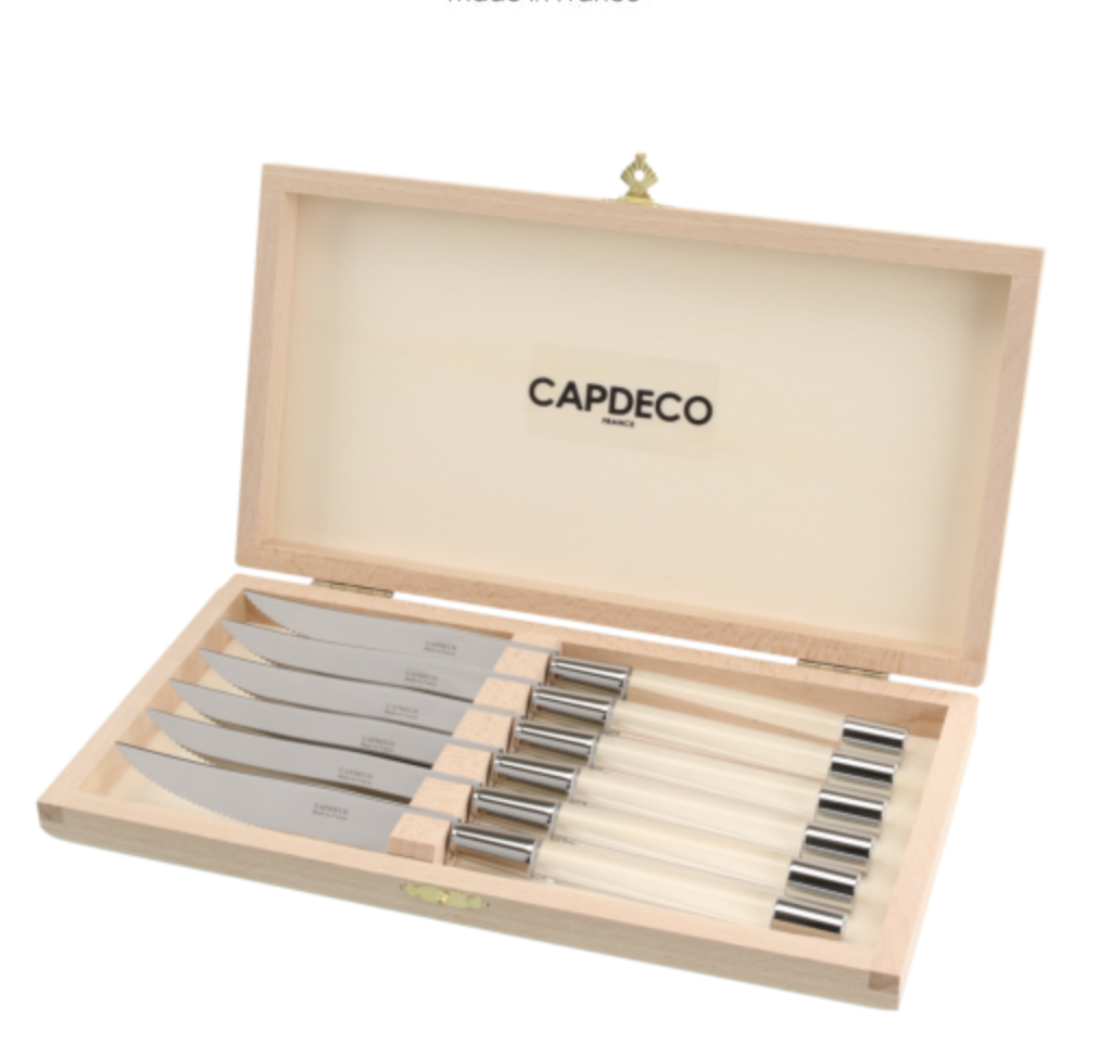 A wooden box with a hinged lid, branded Capdeco inside, is open to display six Zoe Steak Knives made in France, each with a steel blade and light-colored handle, all neatly arranged in individual slots.