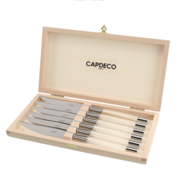A wooden box with a hinged lid, branded Capdeco inside, is open to display six Zoe Steak Knives made in France, each with a steel blade and light-colored handle, all neatly arranged in individual slots.