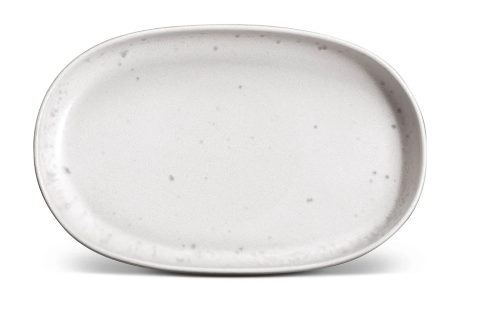 The Terra Stone Dinnerware by L'Objet is an oval, white ceramic plate with gray speckles and a slightly raised rim, beautifully handcrafted and shown on a white background.