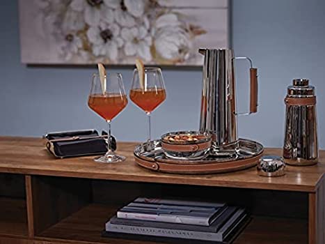 A wooden table holds a metal tray with a pitcher, two glasses of orange drink with pear slices, snacks, the Nambe Tahoe Cocktail Shaker in leather barware, and a clutch. A floral painting hangs on the blue wall behind.
