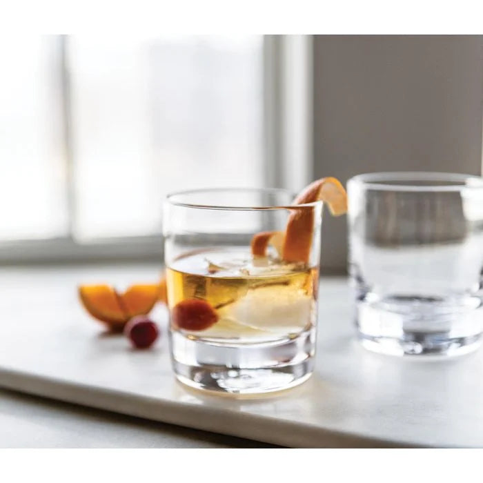 A Simon Pearce Ascutney Double Old Fashioned Set of 2 glass with ice and an orange peel garnish sits on a white surface, its pair nearby. Slices of orange are in the background near a bright window.