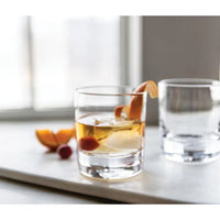 A Simon Pearce Ascutney Double Old Fashioned Set of 2 glass with ice and an orange peel garnish sits on a white surface, its pair nearby. Slices of orange are in the background near a bright window.
