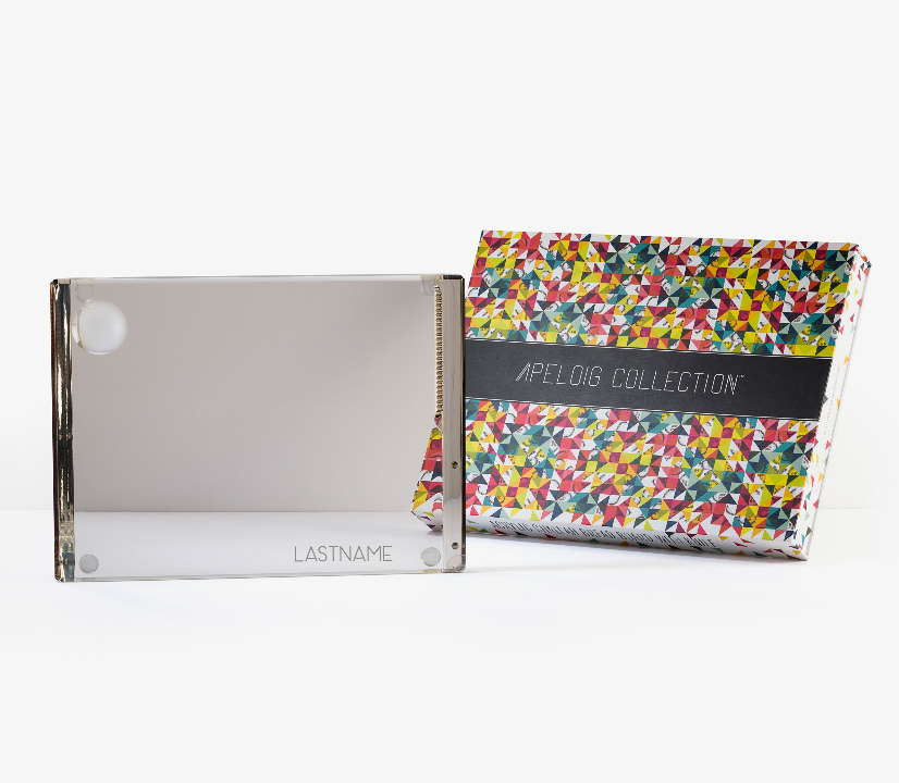 A personalized solid acrylic challah board by Apeloig, custom engraved with LASTNAME, is shown beside a colorful Apeloig box. Both are displayed on a white background.