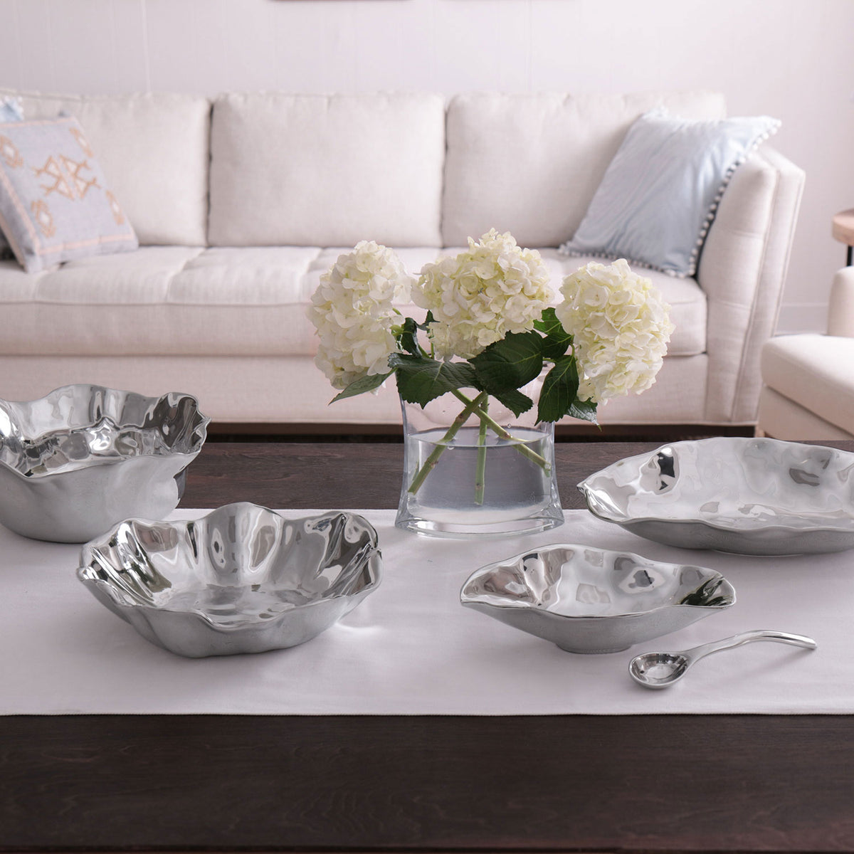 A living room with a beige sofa, light blue pillows, a glass vase of white hydrangeas, and a dark wood table set with five Beatriz Ball Vento Claire Bowls with Spoon on a white runner.