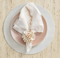 A round white placemat holds a light pink plate and a gold-rimmed white napkin, folded with the Kim Seybert Dahlia Napkin Ring Ivory Set of 4, displayed on a light wooden table.