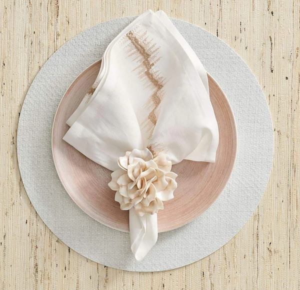 A round white placemat holds a light pink plate and a gold-rimmed white napkin, folded with the Kim Seybert Dahlia Napkin Ring Ivory Set of 4, displayed on a light wooden table.