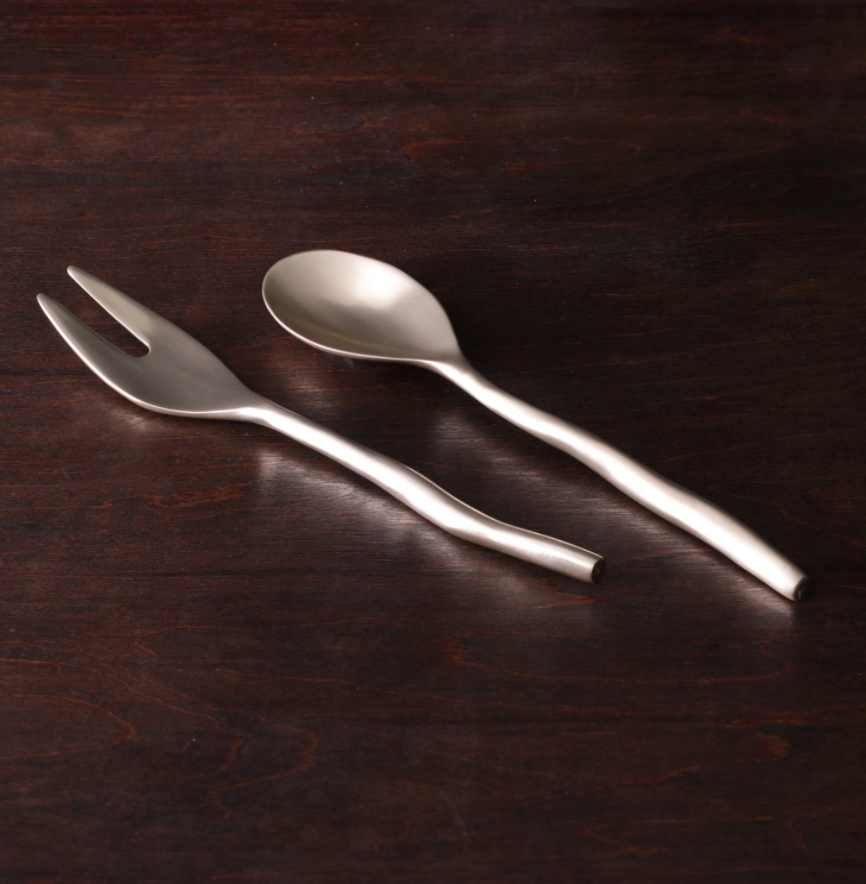 The Sierra Maia Salad Servers by Beatriz Ball feature a metal two-prong fork and matching spoon with wavy handles, placed side by side on dark wood—perfect as eco-friendly serving utensils.