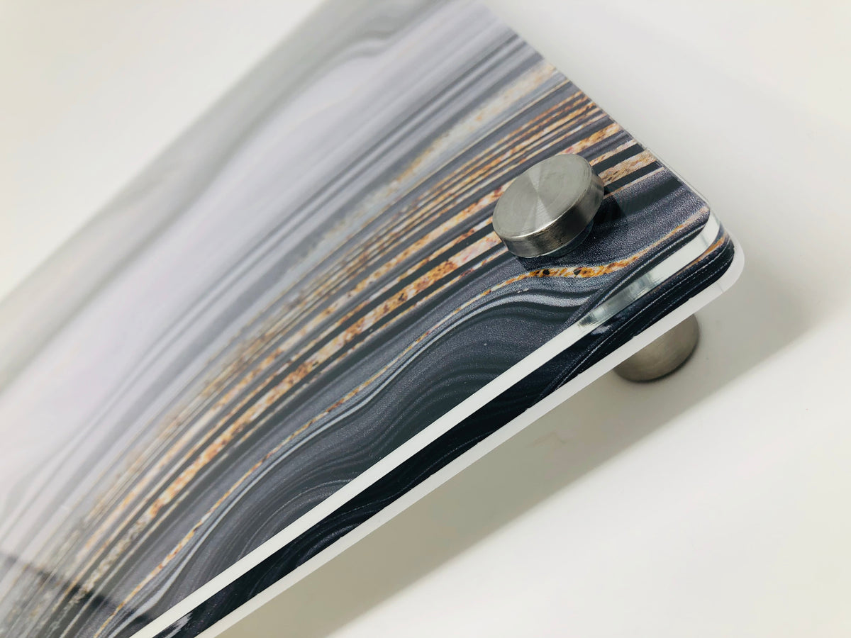 Close-up of a corner of the Raised Black Slice Challah Serving Board by Eye Candy, featuring a marbled striped pattern in gray, white, and brown tones, mounted on a cylindrical metal standoff against a white background.