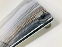 Close-up of a corner of the Raised Black Slice Challah Serving Board by Eye Candy, featuring a marbled striped pattern in gray, white, and brown tones, mounted on a cylindrical metal standoff against a white background.