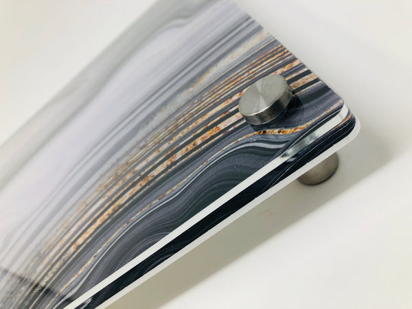 Close-up of a corner of the Raised Black Slice Challah Serving Board by Eye Candy, featuring a marbled striped pattern in gray, white, and brown tones, mounted on a cylindrical metal standoff against a white background.