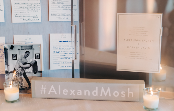A table display featuring handwritten notes, a black-and-white photo of dancing newlyweds, glowing candles, a #AlexandMosh sign, and an Eye Candy Personalized Wedding Invitation Tray holding Alexandra Lauren and Mosheh David's invitation.