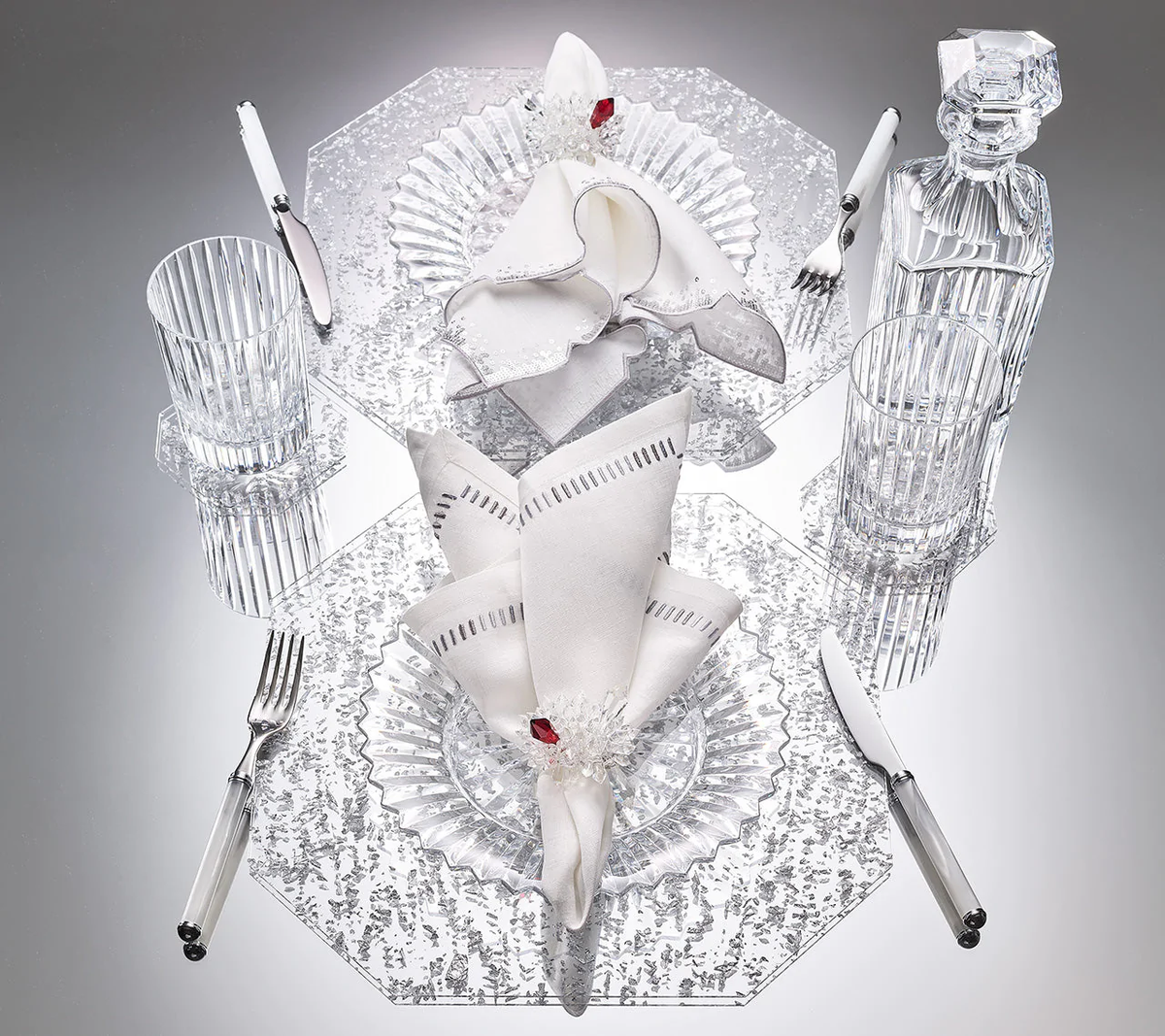 Elegant table setting for two featuring Kim Seybert’s Harmonie Napkin in White, Gray & Silver (set of 4), clear glass plates, cutlery, Baccarat crystal glasses, and a decanter arranged symmetrically on a reflective surface.