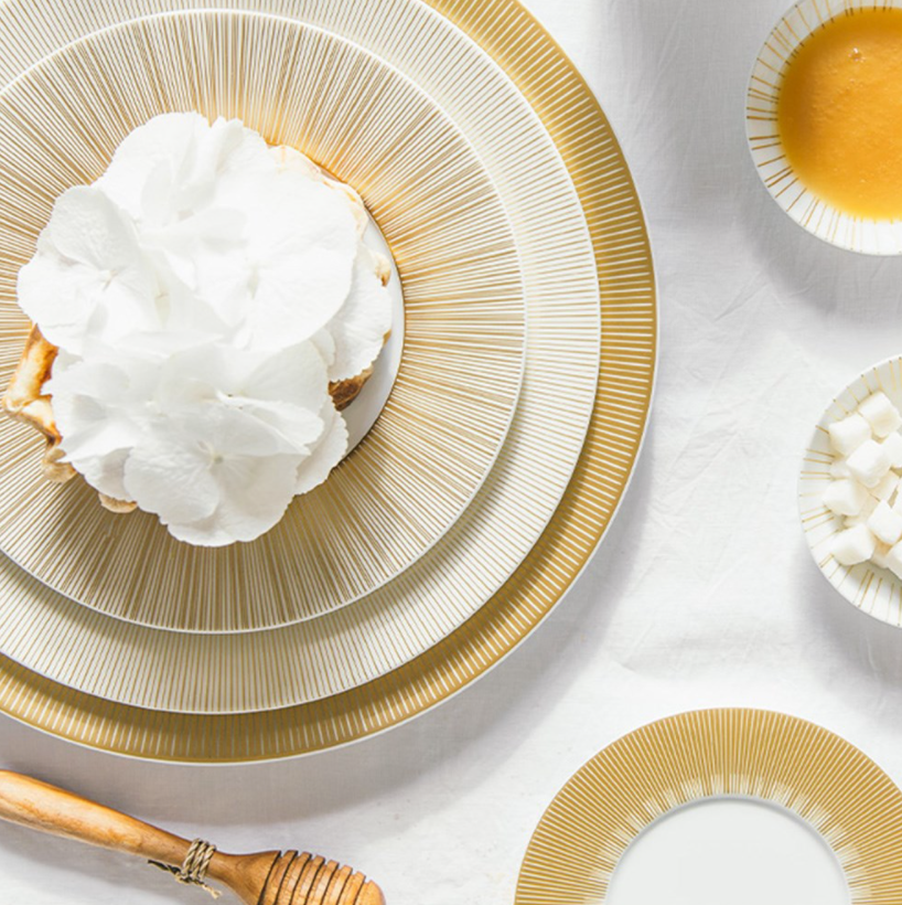 A table set with Bernardaud's Sol Dinnerware and gold-rimmed plates, featuring a dessert garnished with white flowers, a bowl of honey with dipper, and sugar cubes on a white tablecloth.