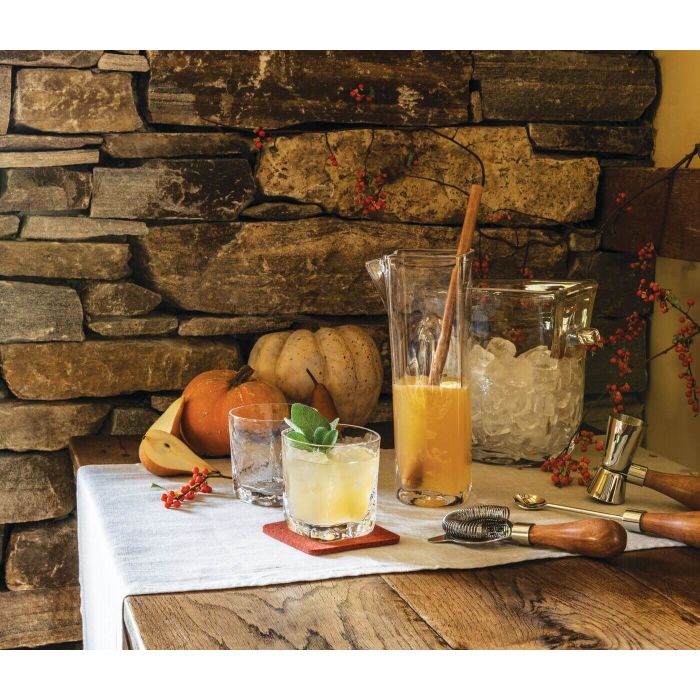 A rustic table with a white cloth displays the Simon Pearce Woodbury Chiller, a pitcher of orange juice with ice and a wooden spoon, two pumpkins, bar tools, and a stone wall adorned with orange berries in the background.