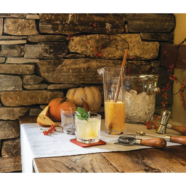 A rustic table with a white cloth displays the Simon Pearce Woodbury Chiller, a pitcher of orange juice with ice and a wooden spoon, two pumpkins, bar tools, and a stone wall adorned with orange berries in the background.
