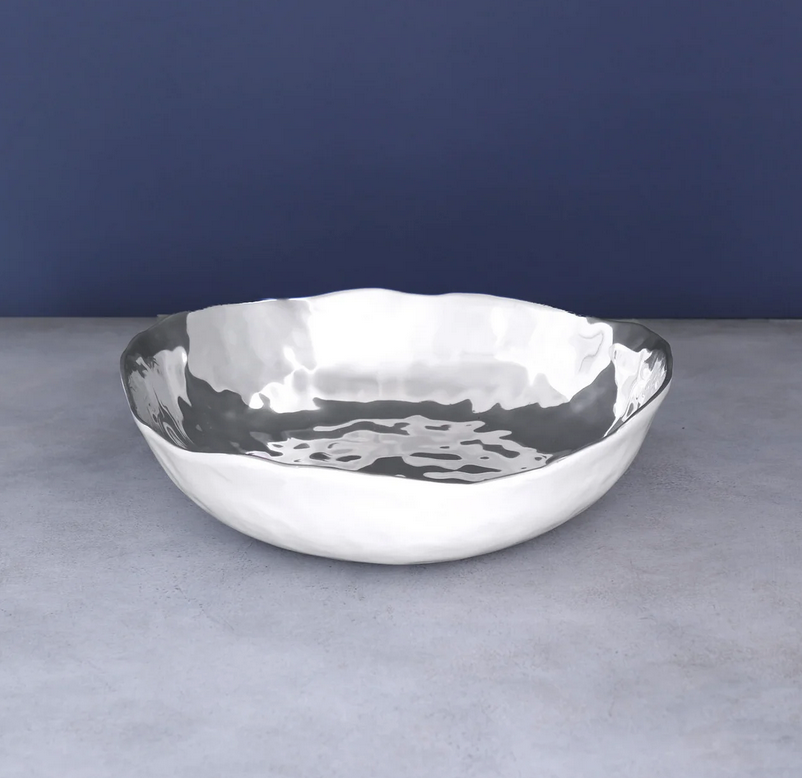 The Beatriz Ball Thanni Osaka Bowl White and Silver Large, with its hammered metal finish and wavy edge, rests on a gray surface against a navy background—ideal as a distinctive housewarming gift or elegant salad bowl.