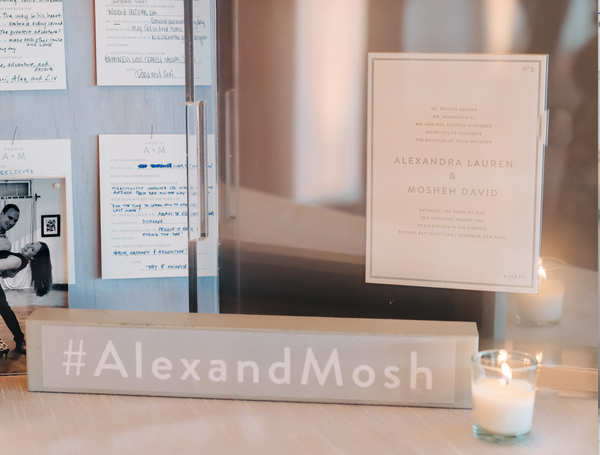 A lightbox sign reads #AlexandMosh on a table with a glowing candle, wedding invitation, displayed notes, and the Eye Candy Personalized Wedding Invitation Tray, setting a soft and elegant celebration scene.