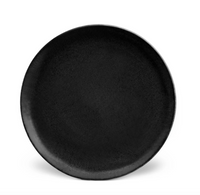 A round, matte black ceramic plate from L’Objet's Terra Iron Dinnerware is shown against a white background. The handcrafted plate has a smooth surface with slightly raised edges, ideal for elegant table settings.