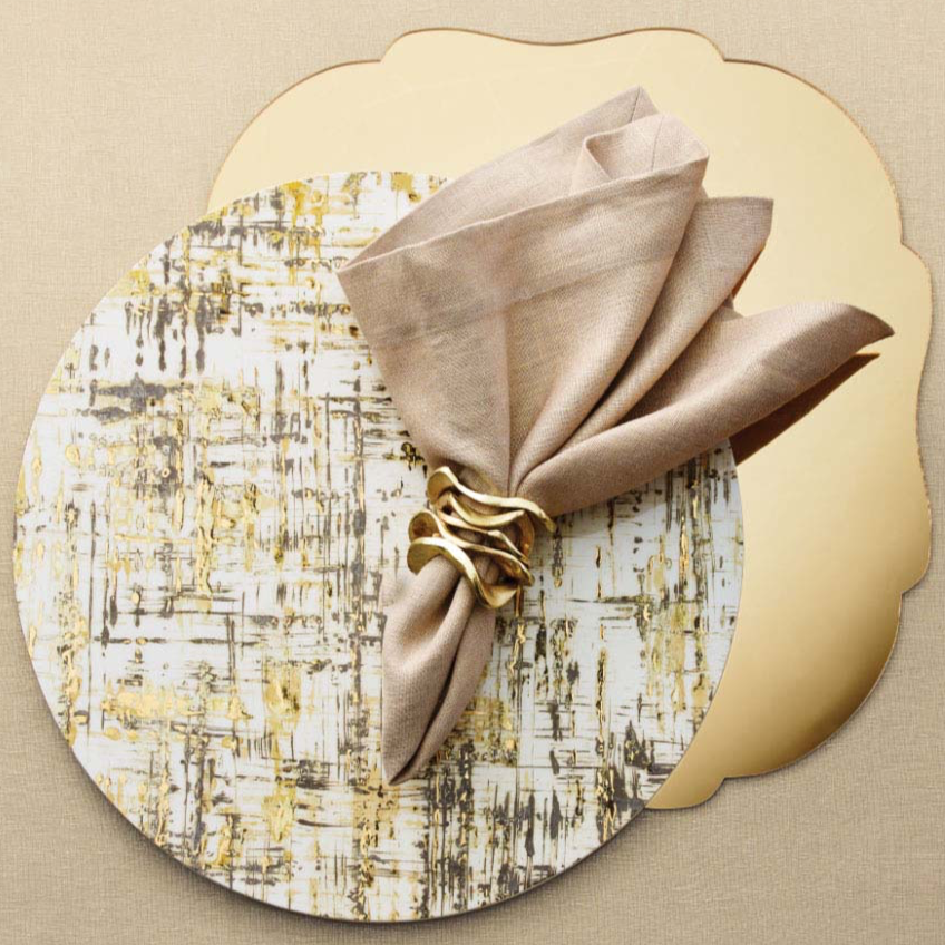 A round plate with an abstract gold and black pattern sits on a gold scalloped placemat. A beige cloth napkin, held by Kim Seybert's Flux Napkin Ring Gold Set of 4, rests on the plate for chic, elegant table decor.