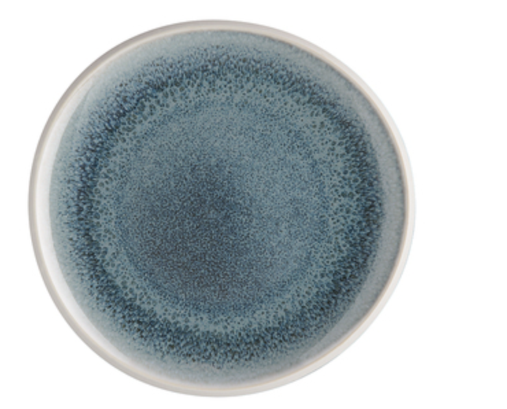 The Rosenthal Junto Aquamarine Dinnerware features a round ceramic plate with a white rim and textured blue gradient center, created with reactive glaze for a unique watercolor effect. This stoneware is dishwasher and microwave safe. Background is white.