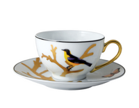 The Bernardaud Aux Oiseaux Dinnerware features a white Limoges porcelain teacup with a gold handle, adorned with a yellow and black bird and gold branches, paired with a matching saucer showcasing butterfly and branch motifs.