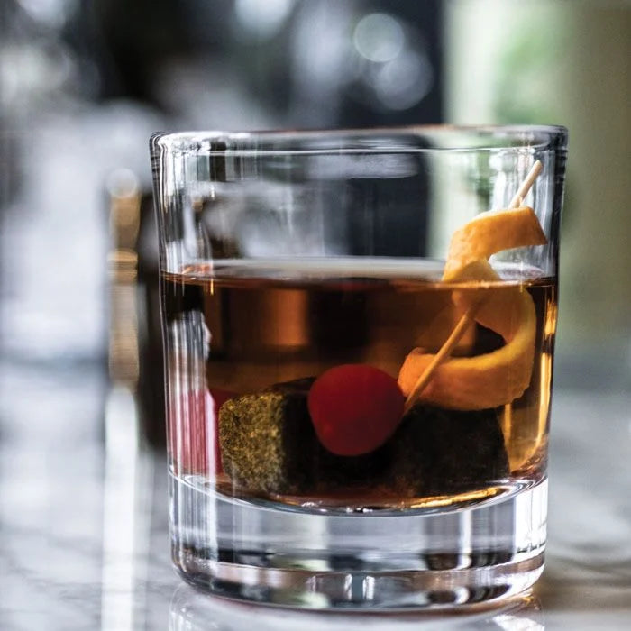 A glass of dark liquor in a Simon Pearce Ascutney Whiskey glass (set of 2) with two whiskey stones, garnished with a cherry and lemon twist on a toothpick, sits on marble with a blurred background.