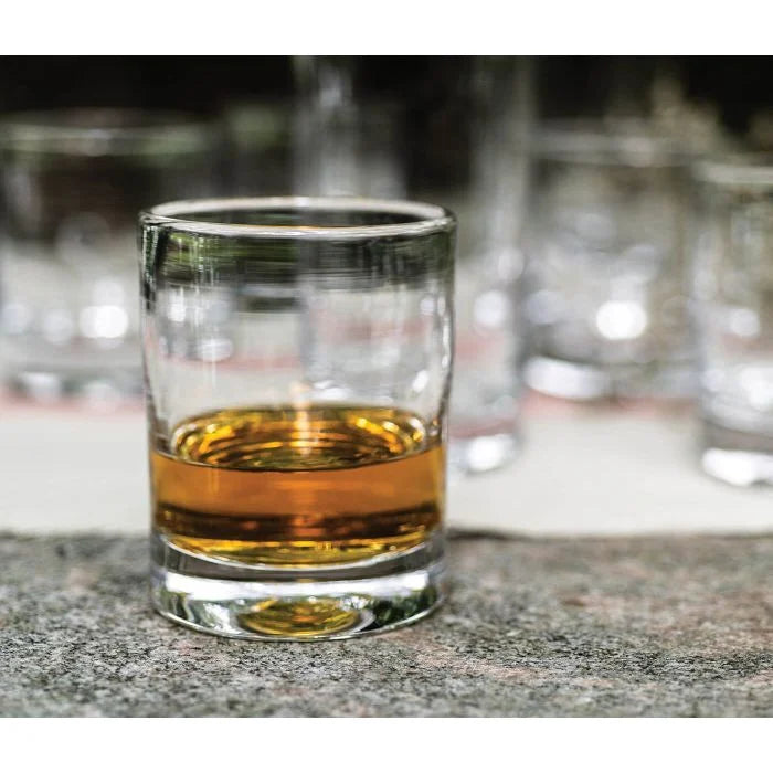 A Simon Pearce Ascutney Whiskey Set of 2 glass, partially filled with amber whiskey, rests on a stone surface while several empty tumblers appear blurred in the background.