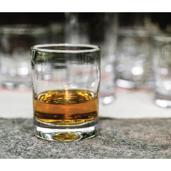 A Simon Pearce Ascutney Whiskey Set of 2 glass, partially filled with amber whiskey, rests on a stone surface while several empty tumblers appear blurred in the background.