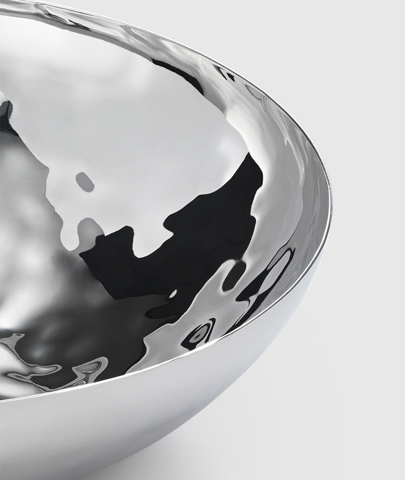 A close-up of the Mary Jurek Luna Serving Bowl 12" shows its polished, reflective stainless steel and smooth curved surfaces against a light grey background. This modern serving bowl is dishwasher safe.