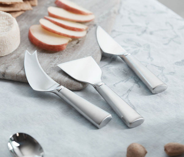 The Simon Pearce Hartland Cheese Knife Set, featuring three stainless steel knives with elegant handles, rests on a light blue floral cloth next to sliced apples, a cheese wheel, and crackers.