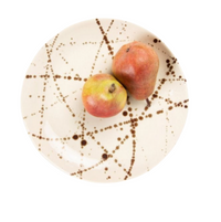 The Delaney Bronze Splatter Dinnerware by Blue Pheasant—a cream stoneware plate with bronze splatters—holds two pears, one small and yellow-green and the other large and reddish-brown, on a white background.