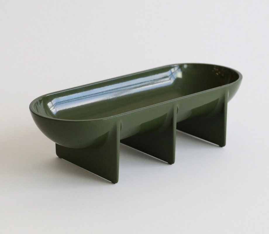 The Standing Bowl Oval by FS Objects is a modern fruit bowl with a dark green, oval design, elevated by three vertical supports. Made of die cast aluminum, this centerpiece adds style and contrast to any light background.