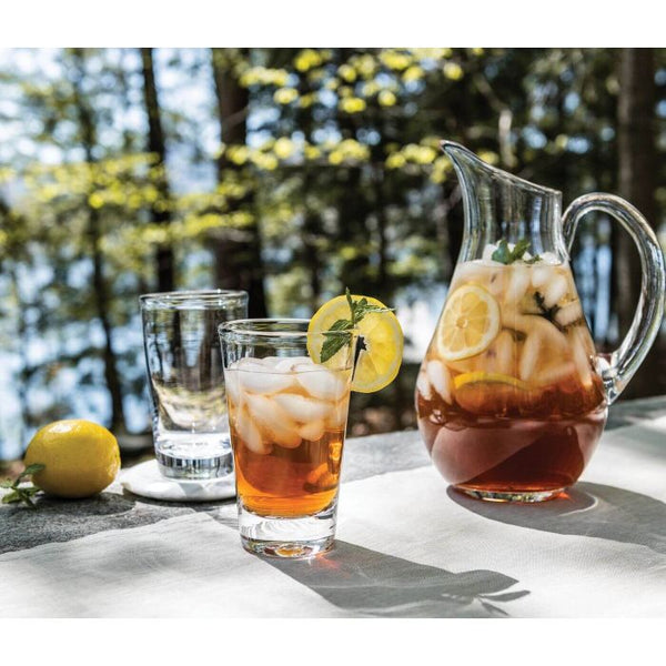A Simon Pearce Ascutney Pint filled with iced tea, lemon slices, and mint sits beside an empty pint, a whole lemon, and sunlight filtering through trees for a perfect summer scene.