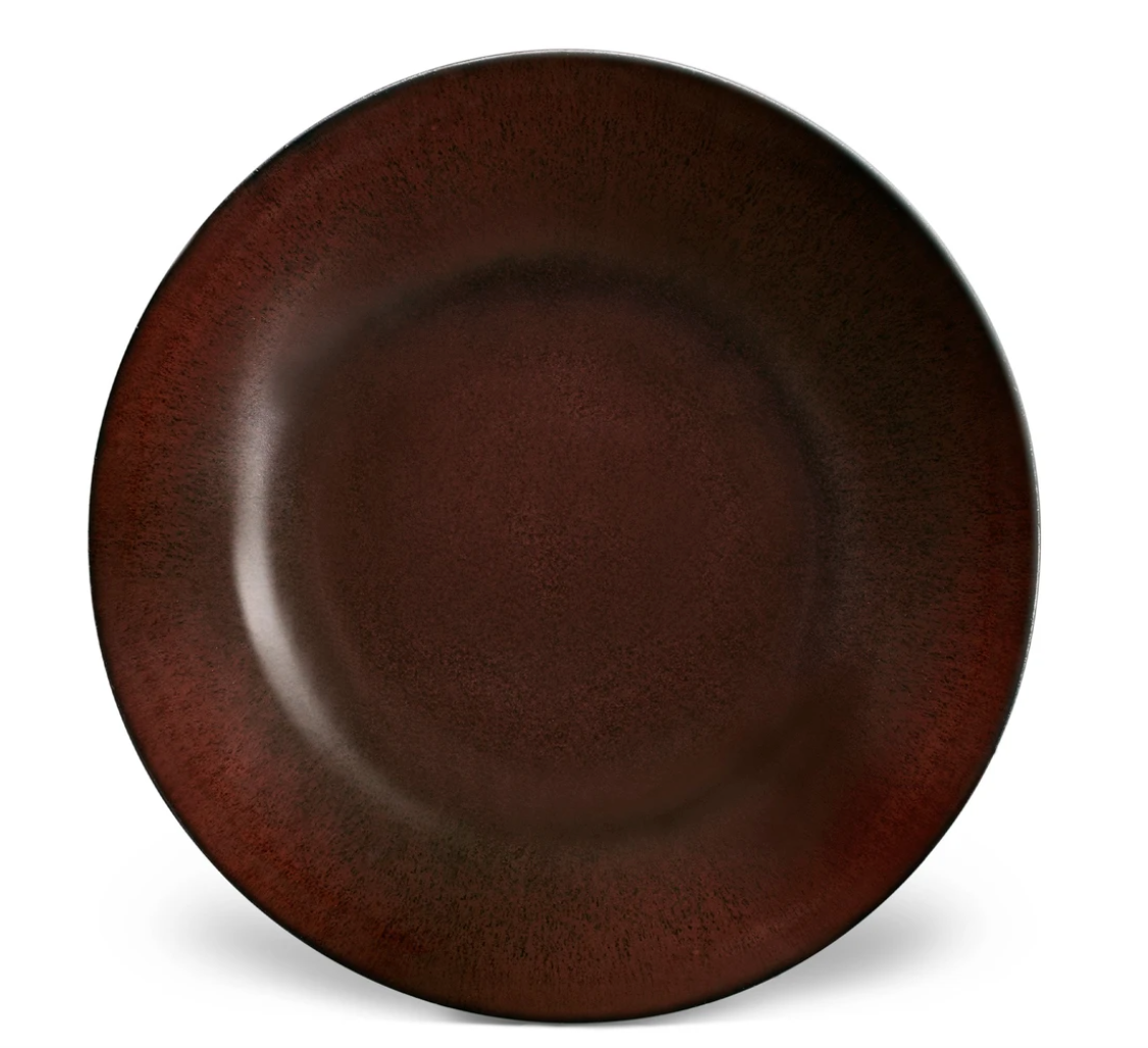 The Terra Wine Dinnerware by L'Objet is a round ceramic plate with a deep reddish-brown hue, darker at the rim and lighter in the center, set against a white background.