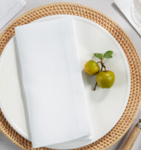 A folded Simple Linen Napkin White from Saro Trading rests on a white plate atop a woven placemat, accompanied by two small green fruits with leaves.