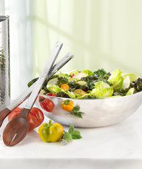 The Mary Jurek Luna Serving Bowl 12" is filled with leafy green salad and vegetables, placed on a marble surface beside wooden salad tongs, fresh tomatoes, and herbs.